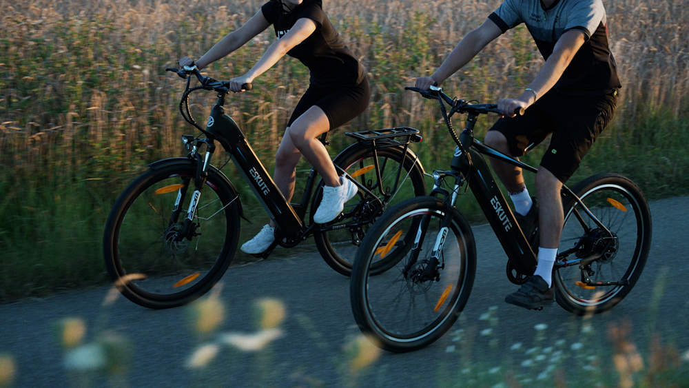 a man and a woman are pedalling their e-bikes on the path surrounded by fields