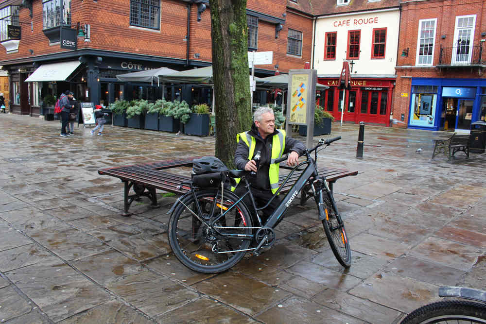 a old man sits on the bench for a drink behind his e-bike